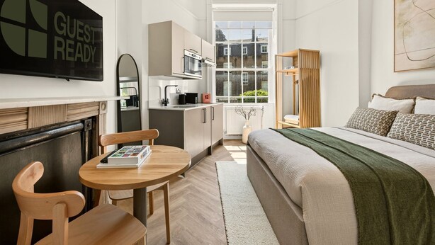 Ett kompakt hotellrum med en säng, ett litet bord med stolar, ett litet kök och ett stort fönster.