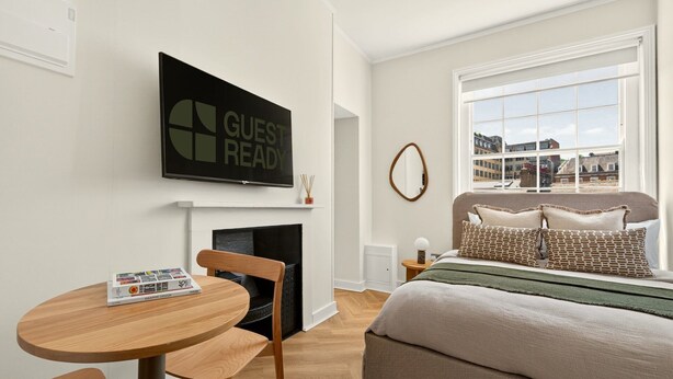 Una habitación de hotel moderna con chimenea, una cama con almohadas estampadas, una mesa redonda de madera con una revista y un televisor mostrando 'GUEST READY'.