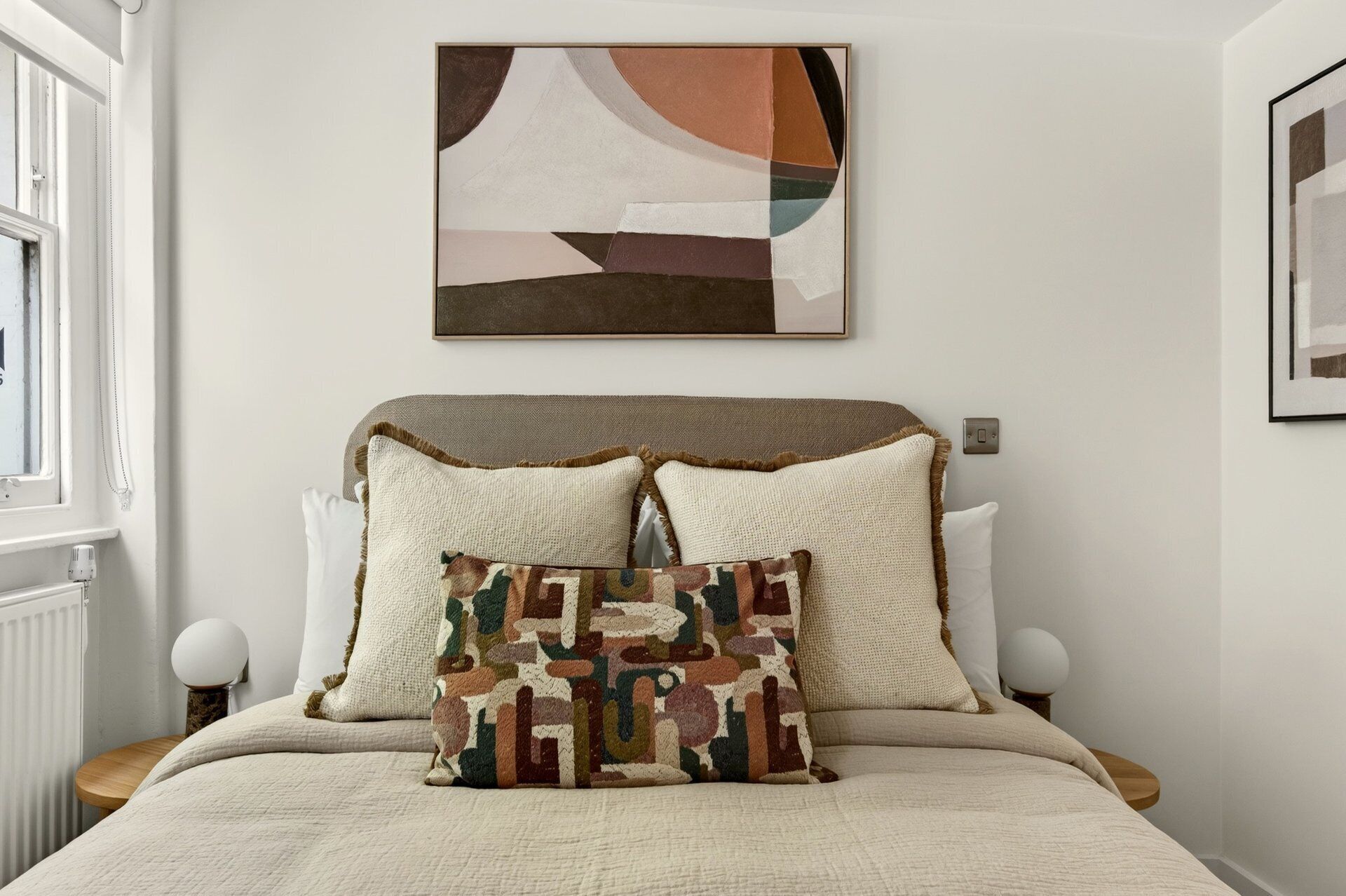 A bedroom with a bed, two bedside tables, a wall-mounted abstract painting, and a window.