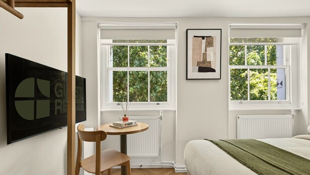 Een slaapkamer met een bed, een stoel, een klein tafeltje, twee ramen, een radiator en een lijst met een foto aan de muur.