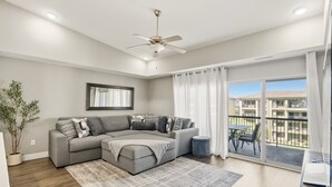 Un salon moderne avec un canapé d’angle, un ventilateur de plafond, une grande fenêtre avec des rideaux et une vue sur un quartier résidentiel.