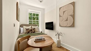 A bedroom with a bed, a bedside table with a book, a chair, a window, and a wall-mounted TV. - GuestReady - Bright Double Room near Euston (London)