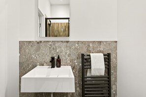 A bathroom with a white sink, a mirror, a towel rack with a white towel, and a shower area with a wooden door. - GuestReady - Modern Double Room near King's Cross (London)