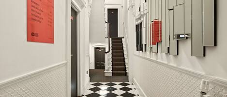 Un couloir d’hôtel avec un sol à carreaux, un escalier et un panneau fixé au mur indiquant les numéros de chambre et les étages.