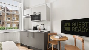 A modern kitchen with a built-in oven and microwave, a small dining area with two chairs, and a window with a view of buildings. - GuestReady - Bright Double En-Suite near Euston (London)
