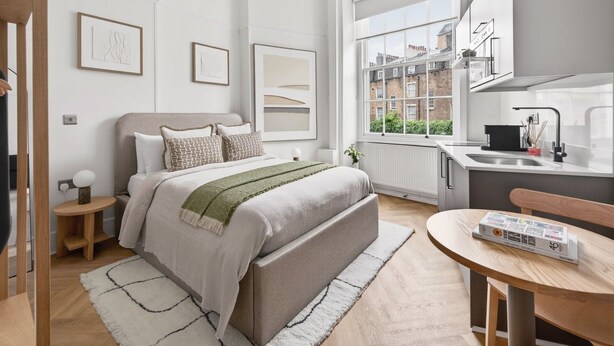 A modern bedroom with a large bed, bedside tables, a view of the city through a window, and a kitchen area with a sink and countertop. - GuestReady - Bright Double En-Suite near Euston (London)