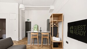 Een moderne hotelkamer met een houten visgraatvloer, een grijze bank, twee houten stoelen, een kleine kitchenette en een bordje met de tekst 'GUEST READY' aan de muur.