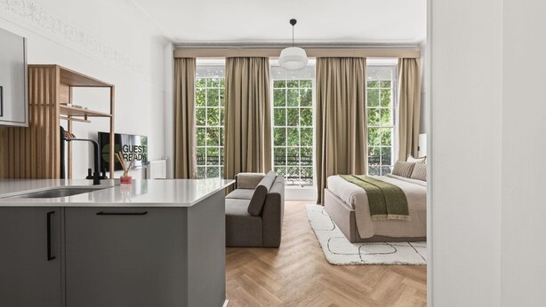 Een moderne hotelkamer met een kitchenette, een bank, een bed en grote ramen met gordijnen.