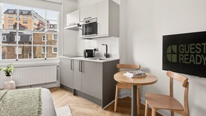 Una cocina moderna con microondas, hervidor y lavamanos, una zona de comedor con mesa y sillas, y vistas a edificios a través de una ventana.