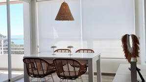 A modern dining area with a white table and chairs, a hanging woven light fixture, and large windows offering a view. - Beautiful apartment overlooking the Mediterranean. (Villajoyosa/Vila Joiosa, La)