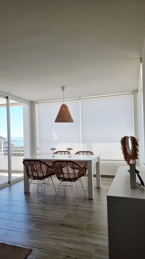A modern dining area with a white table and chairs, a hanging woven light fixture, and large windows offering a view. - Beautiful apartment overlooking the Mediterranean. (Villajoyosa/Vila Joiosa, La)