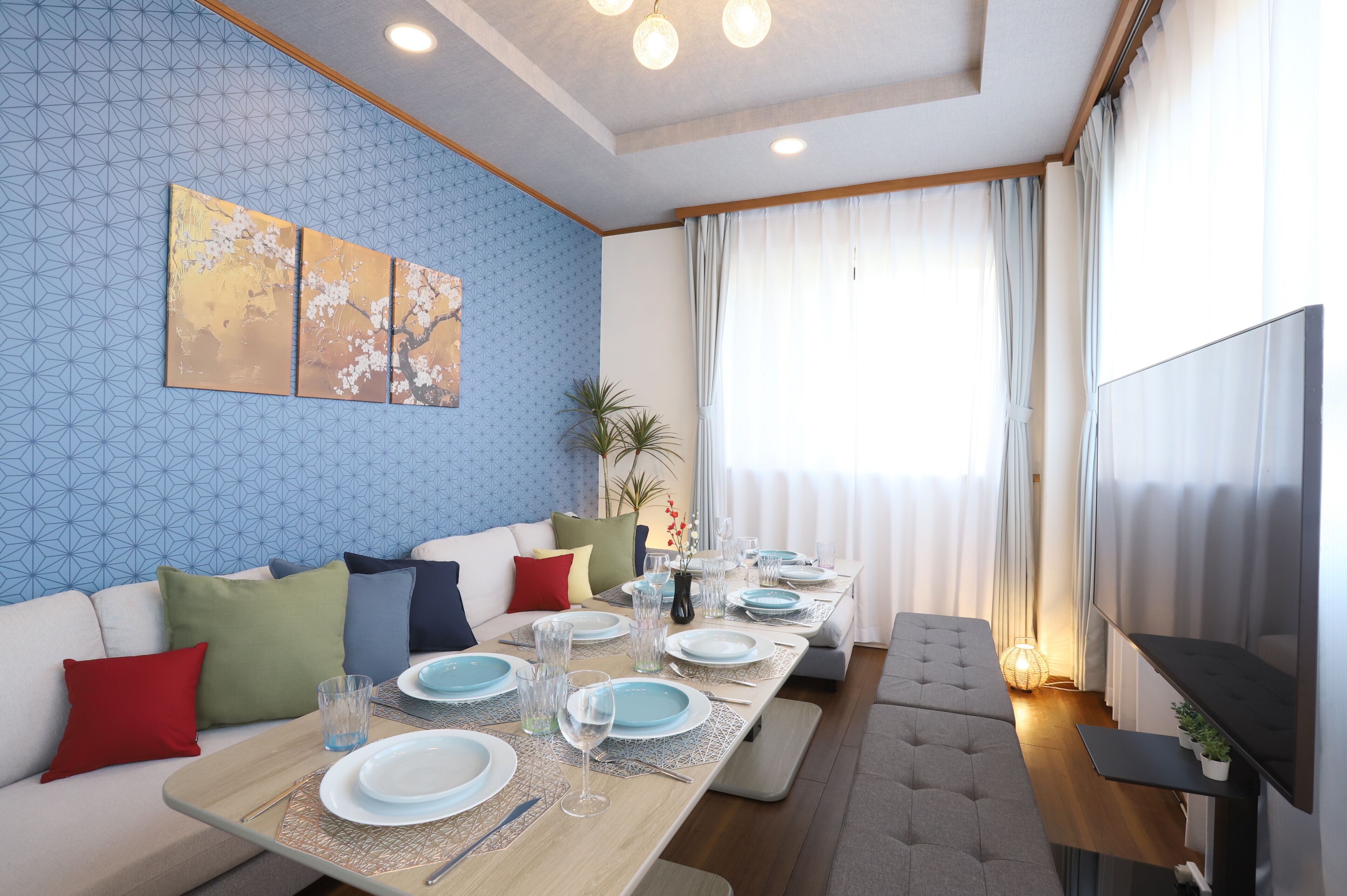 A dining area with a long table set for six, a sofa with cushions, a TV, and a window with curtains.