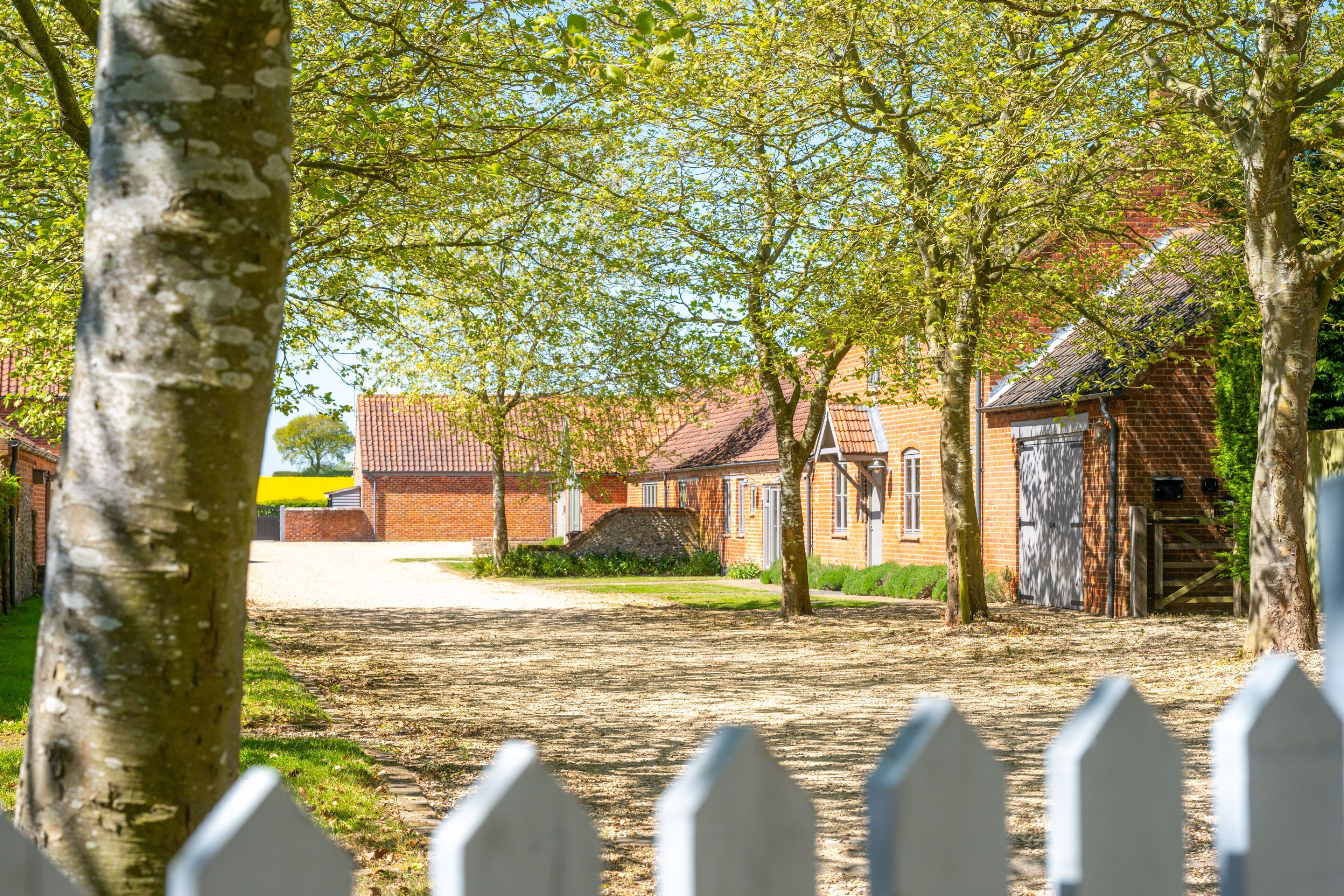 A rural setting with a brick building, a white picket fence, and trees.