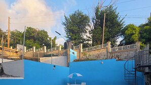 A swimming pool with lounge chairs and a table, surrounded by a blue wall and a fence. - Barefoot Apartments (Roseau)