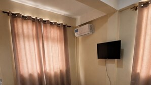 A single bed with a mattress, pillows, an air conditioning unit, a television mounted on the wall, and a standing fan. - Barefoot Apartments (Roseau)