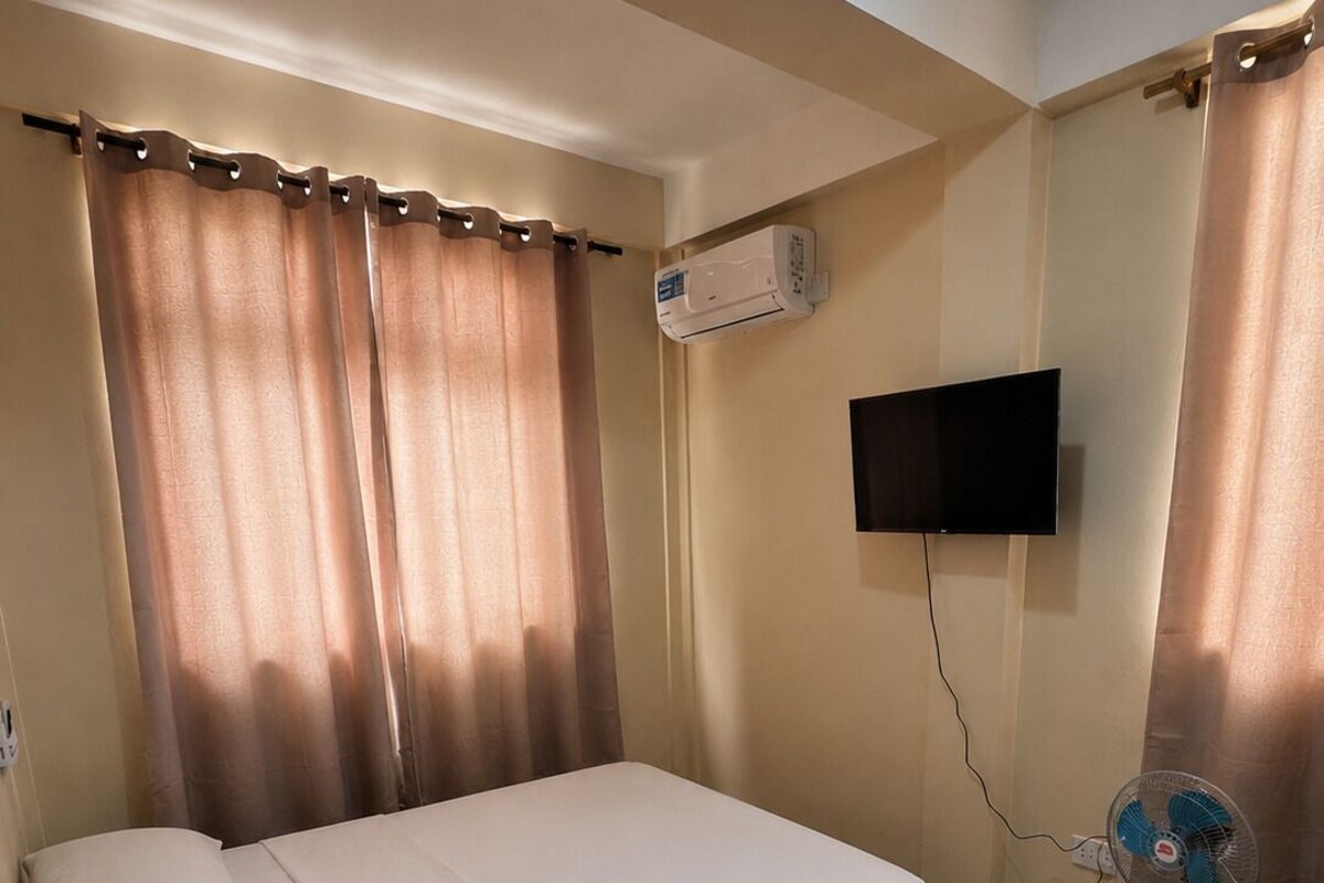 Una cama individual con colchón, almohadas, un aire acondicionado, un televisor fijado a la pared y un ventilador de pie.