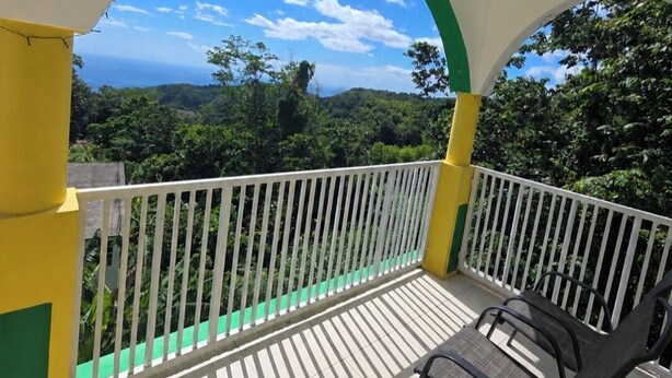 A balcony with a white railing, a view of a green landscape, and a clear sky. - Barefoot Apartments (Roseau)
