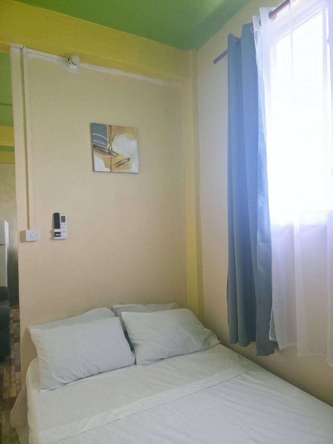 Una habitación con una cama, aire acondicionado de pared, un cuadro pequeño y una ventana con cortinas.