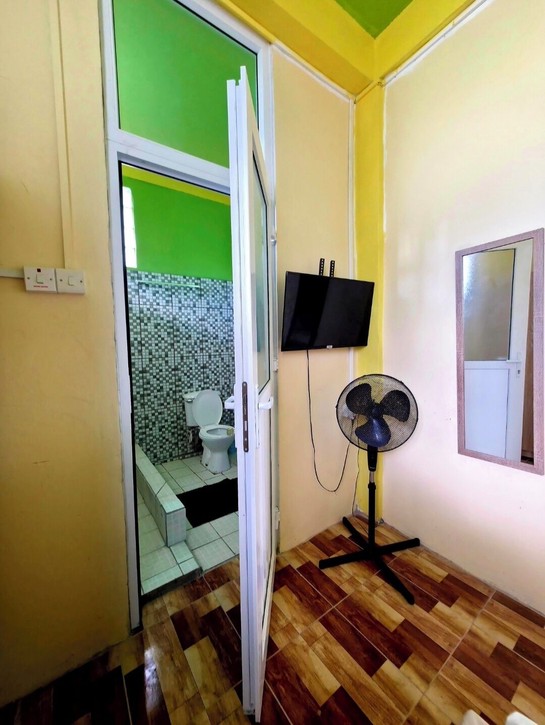 A bathroom with a toilet, a shower area, a fan, and a television mounted on the wall.