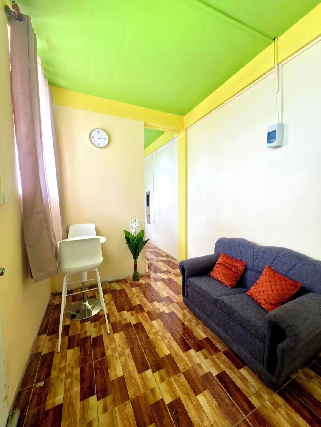 A room with a checkered wooden floor, a grey sofa with orange pillows, a white chair, a clock on the wall, and a green ceiling.