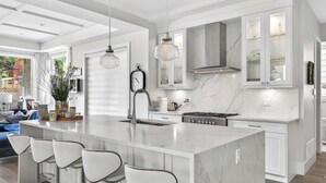 A modern kitchen with a large island, bar stools, a clock, and a stove. - Luxury 4BR | Steps to Beach, Hot Tub & Theatre (Surrey)