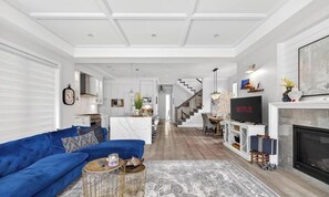 A modern living room with a blue sofa, a fireplace, a TV, and a staircase leading to another level. - Luxury 4BR | Steps to Beach, Hot Tub & Theatre (Surrey)