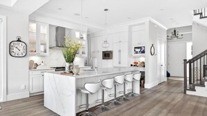 A modern kitchen with white cabinetry, a marble island, bar stools, and a large clock on the wall. - Luxury 4BR | Steps to Beach, Hot Tub & Theatre (Surrey)