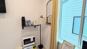 A hotel room with a flat-screen TV, a microwave, a coffee maker, a lamp, a framed welcome sign, and a window with curtains.