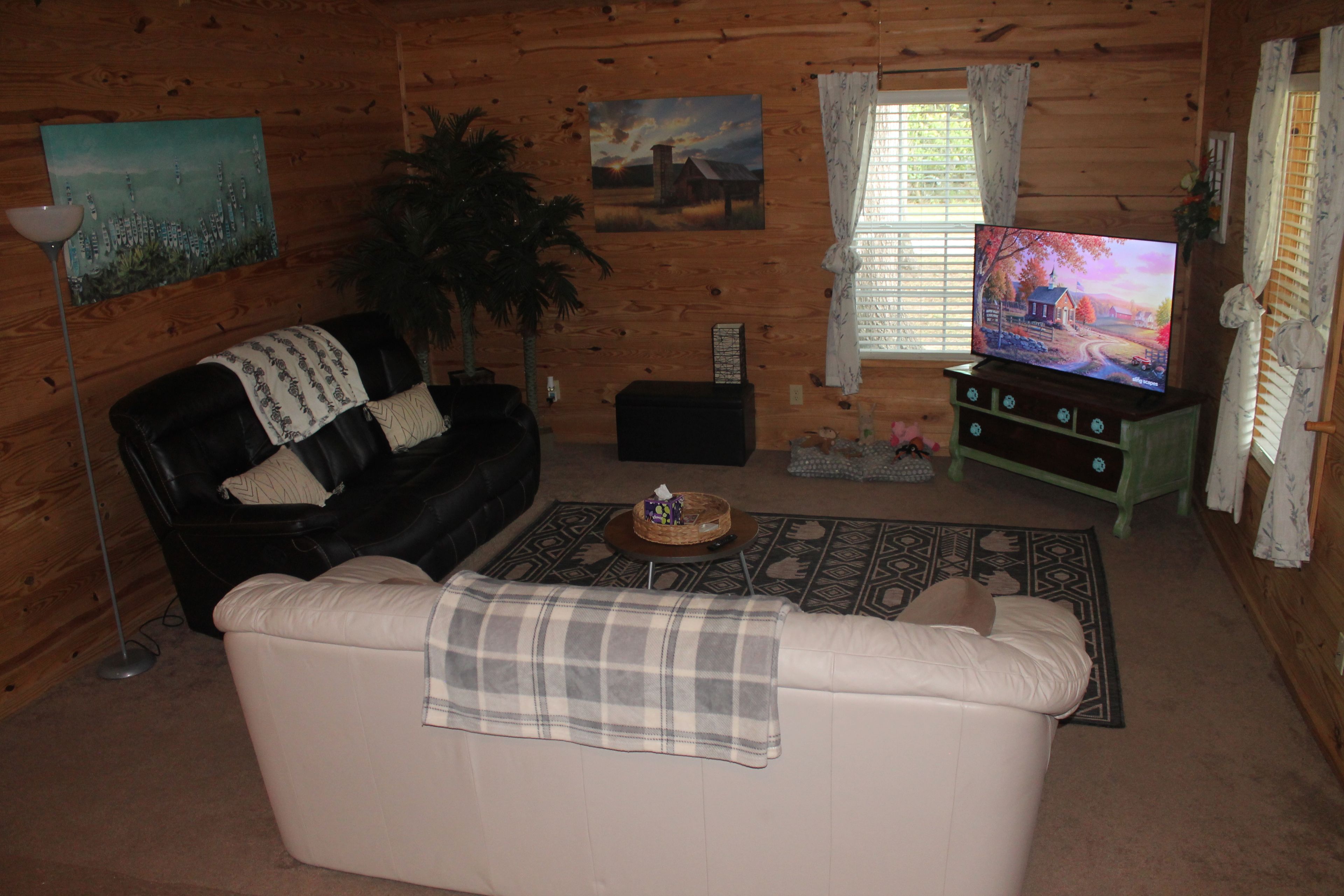 A living room with a black leather sofa, a white sofa, a coffee table, a television, and a large window with curtains.