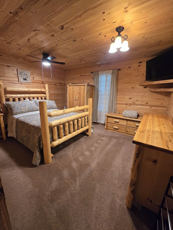 A wooden cabin interior with a bed, a desk, a television, and a ceiling fan.