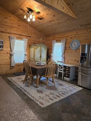 A wooden interior with a dining table, chairs, a clock, and a refrigerator.