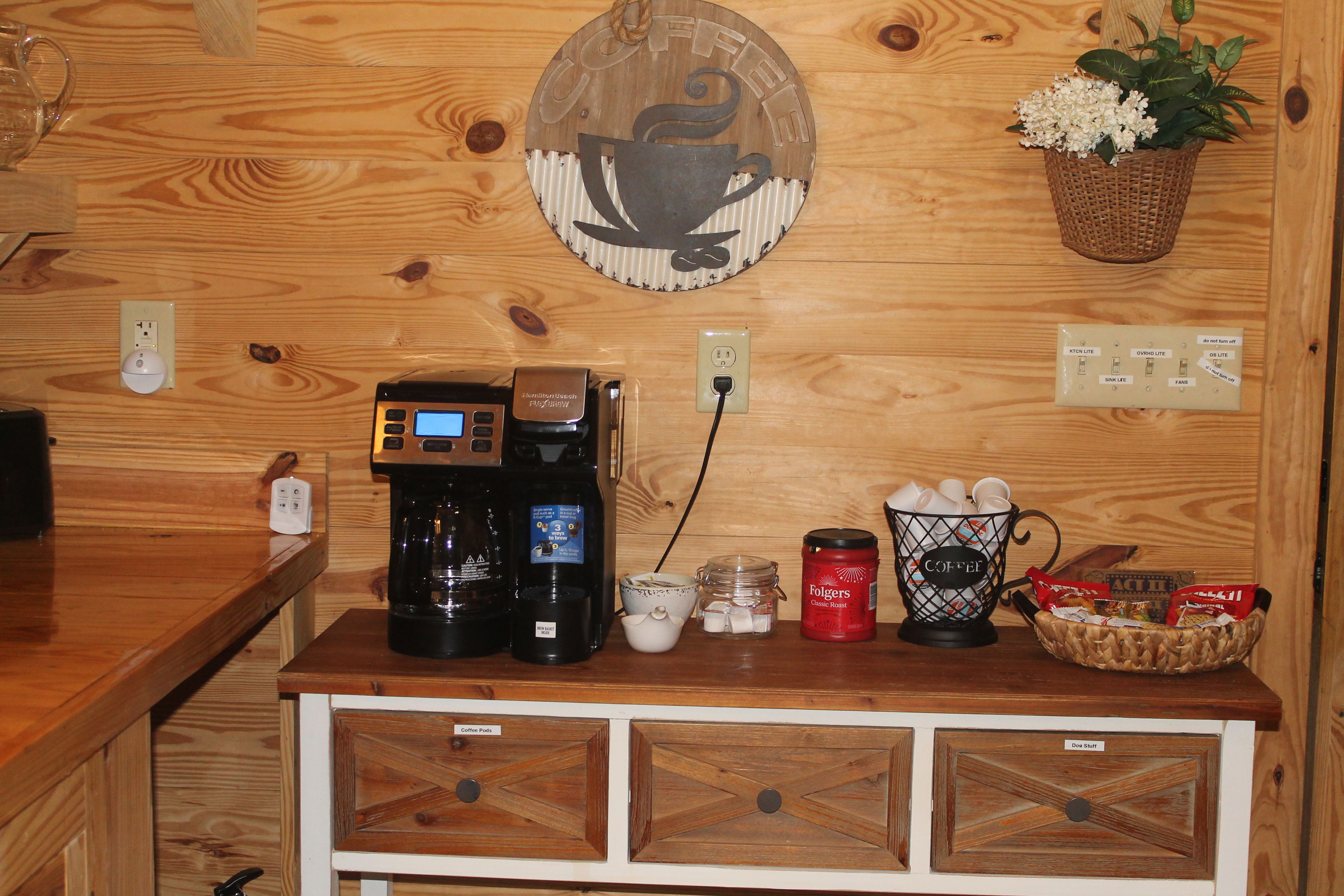 A wooden wall with a coffee machine, a basket of coffee pods, a mug, and a container of Folgers coffee.