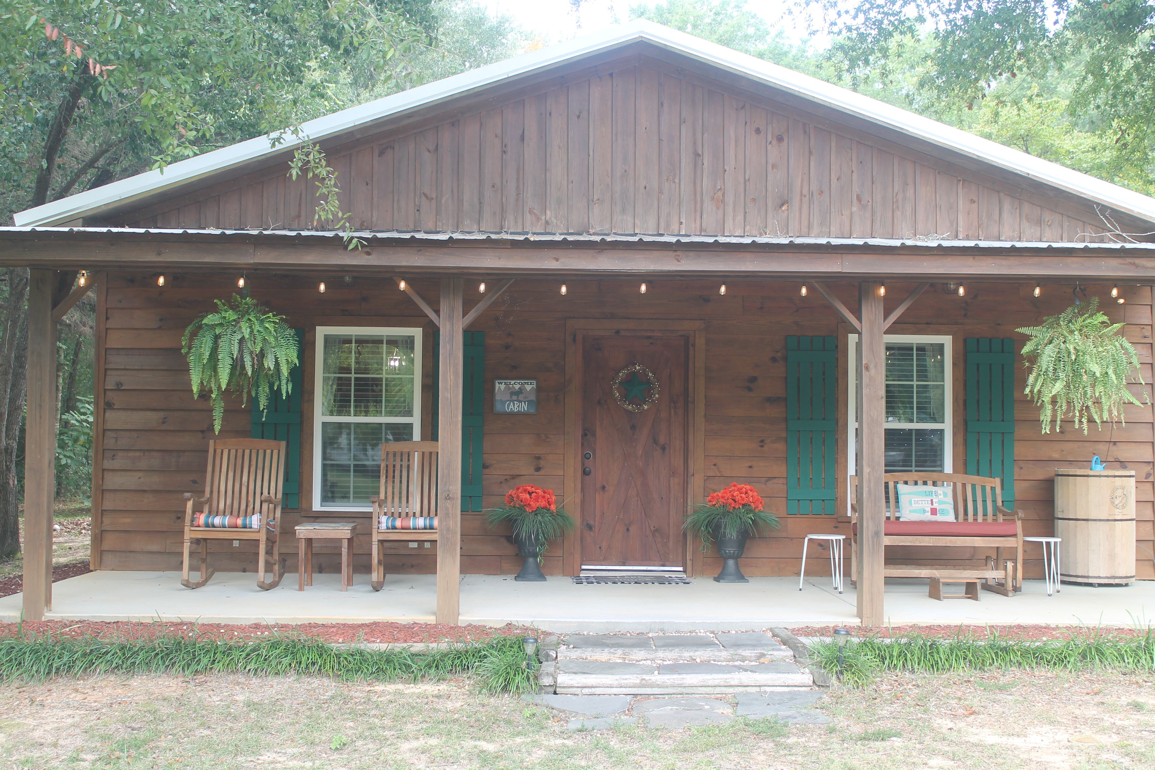A wooden cabin with a porch, two chairs, a bench, and hanging plants.