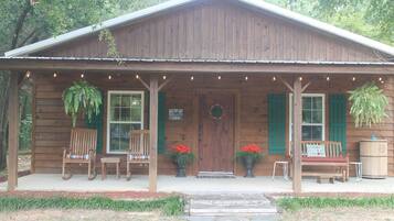 A wooden cabin with a porch, two chairs, a bench, and hanging plants.