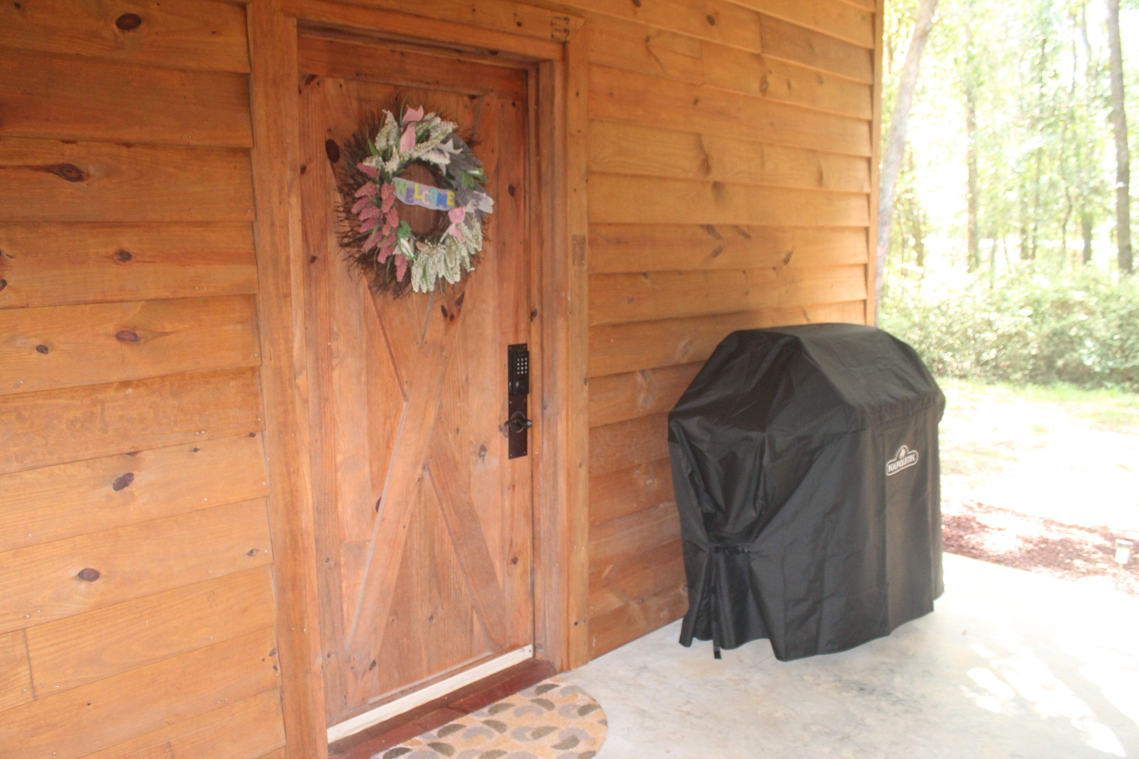 A wooden cabin entrance with a door, a welcome wreath, and a covered grill.