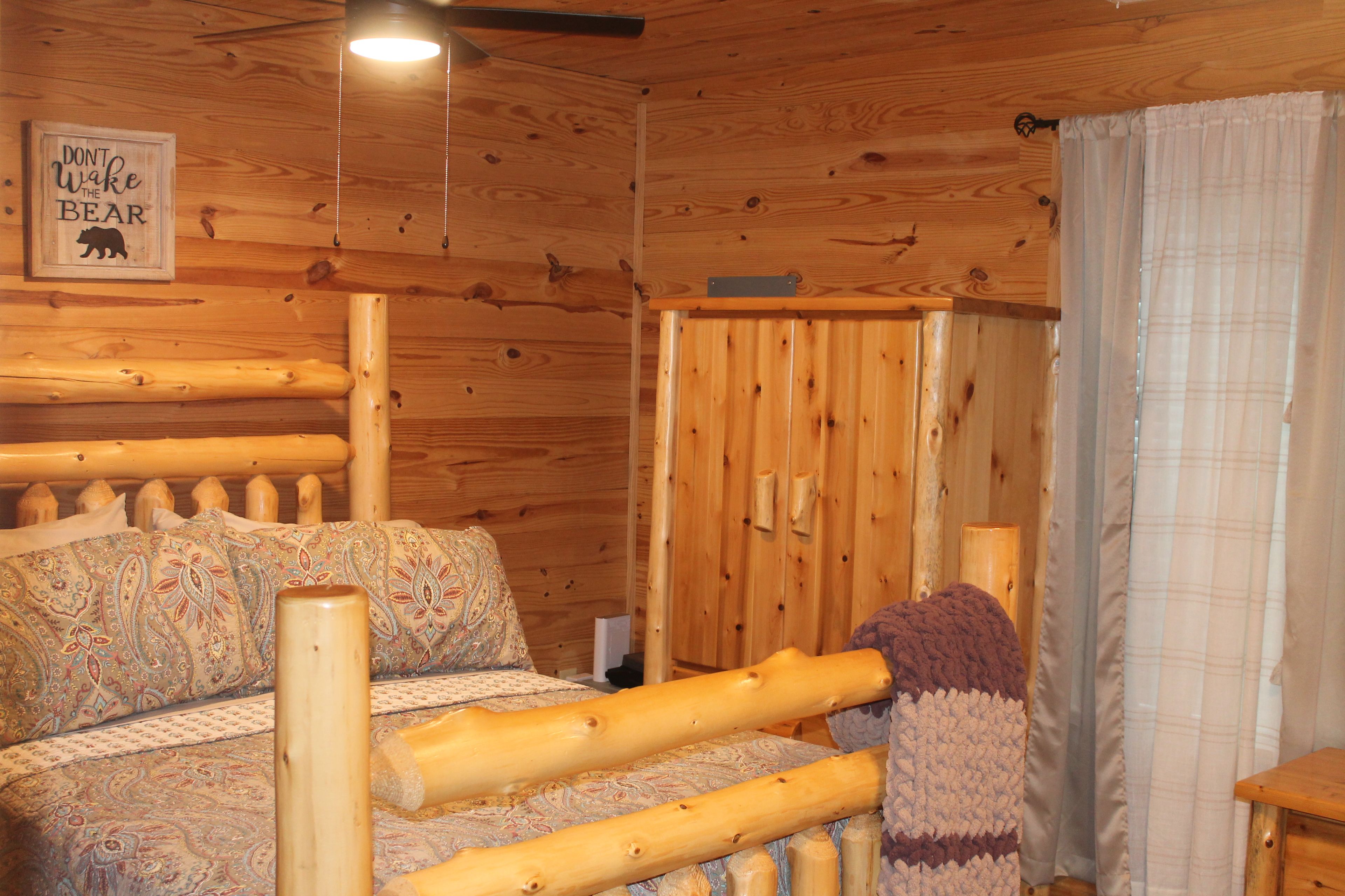 A wooden cabin interior with a bed, a wall-mounted sign, and a window with curtains.