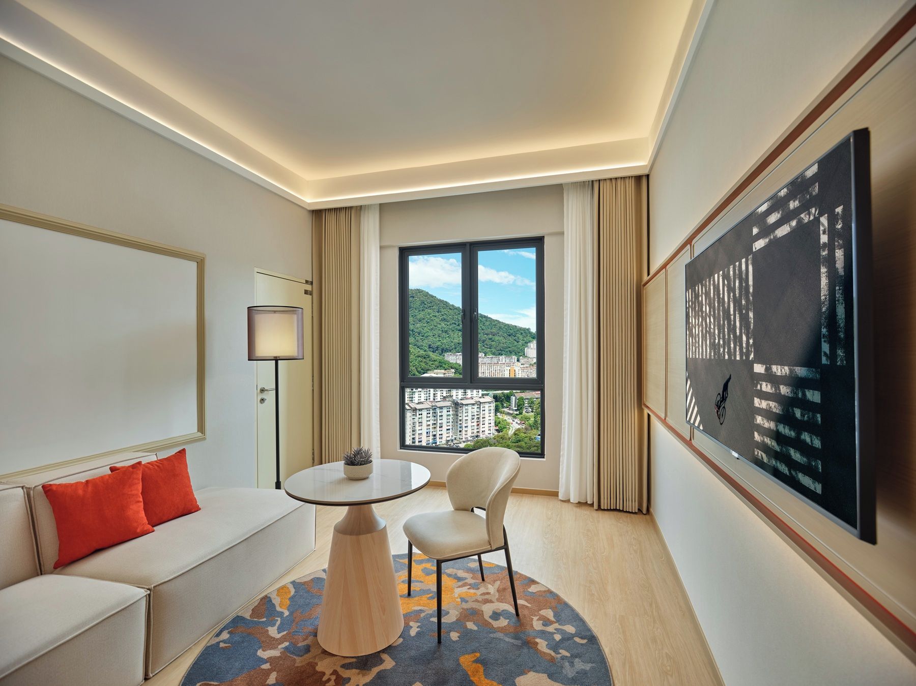 A hotel room with a sofa, a table, a chair, a large window with a view of buildings, and a painting on the wall.
