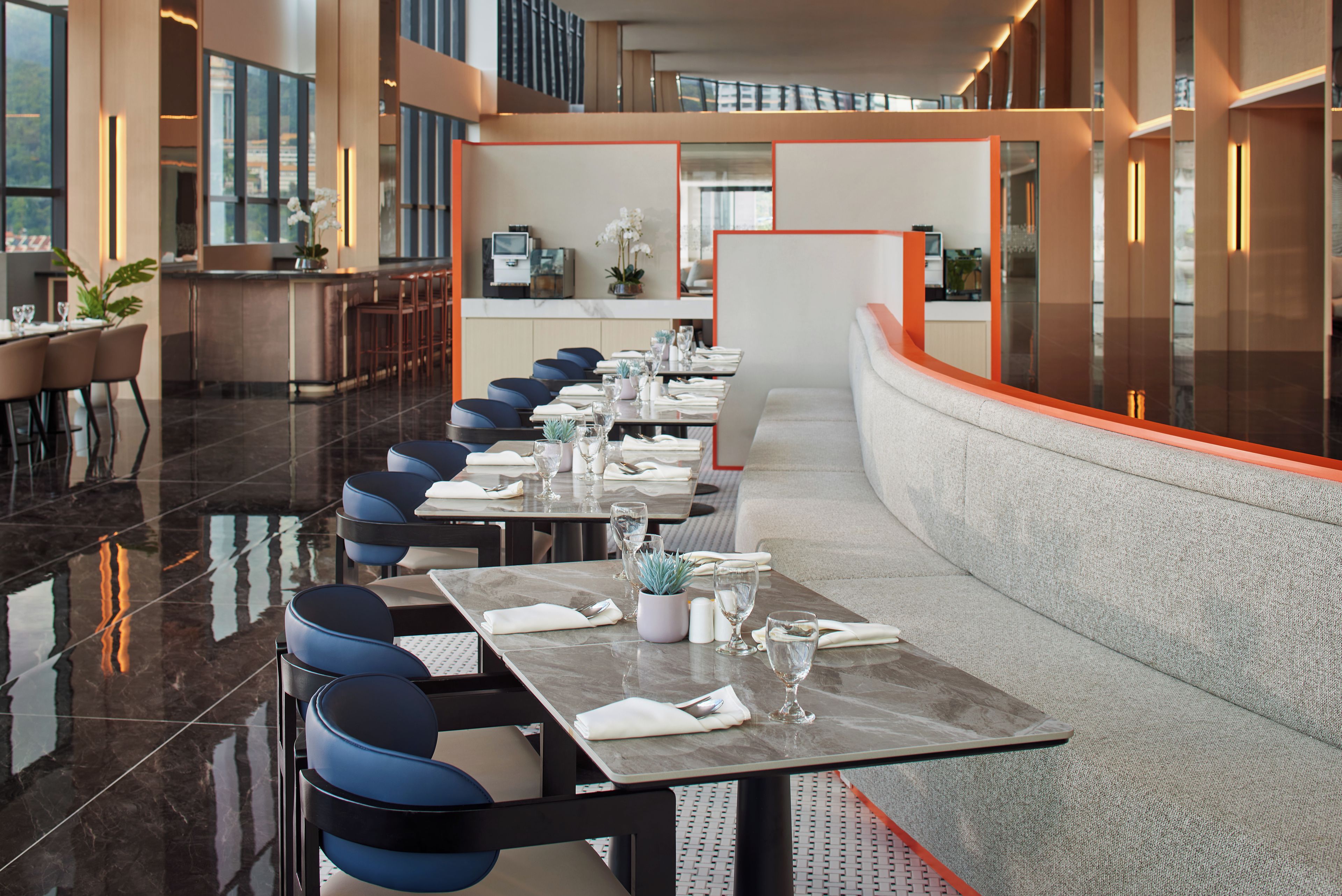 A modern hotel dining area with a long table set for a meal, surrounded by blue chairs.