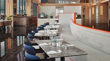 A modern hotel dining area with a long table set for a meal, surrounded by blue chairs.