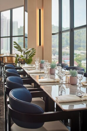 A dining area with a long table set for a meal, large windows, and a view of buildings.