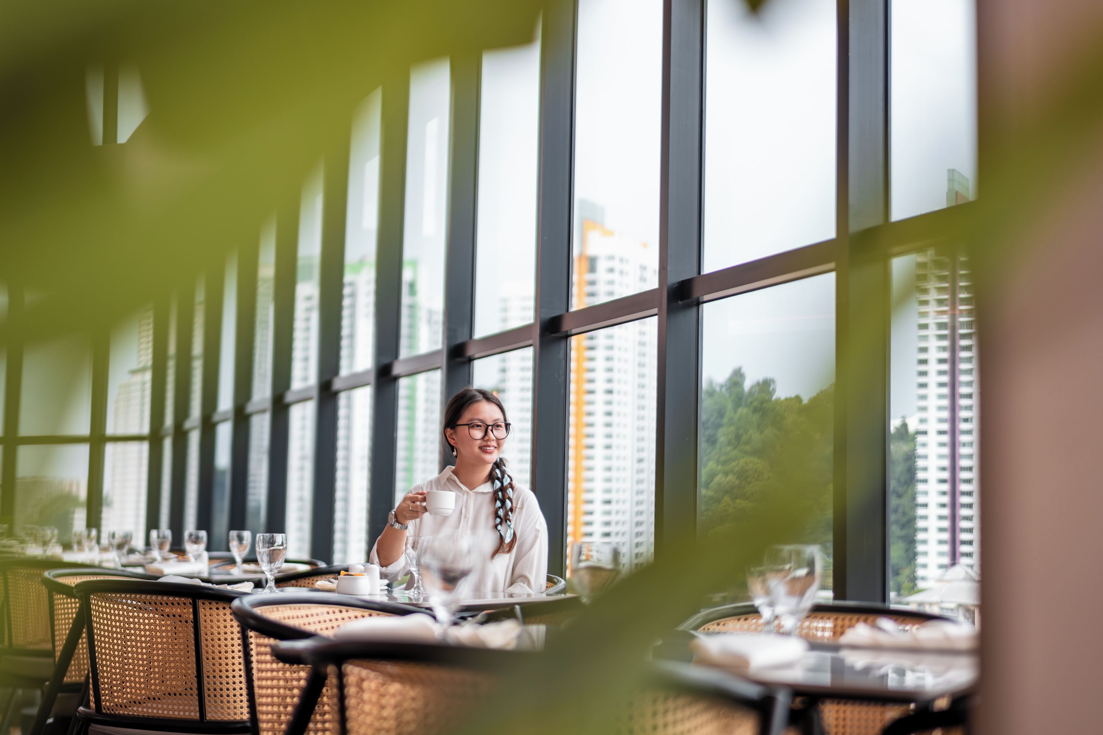 Un restaurante con amplios ventanales, mesas dispuestas con vasos y cubiertos, y vista a un paisaje urbano.