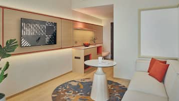 A modern hotel room with a sofa, a round table, a wall-mounted TV, and a kitchen area.