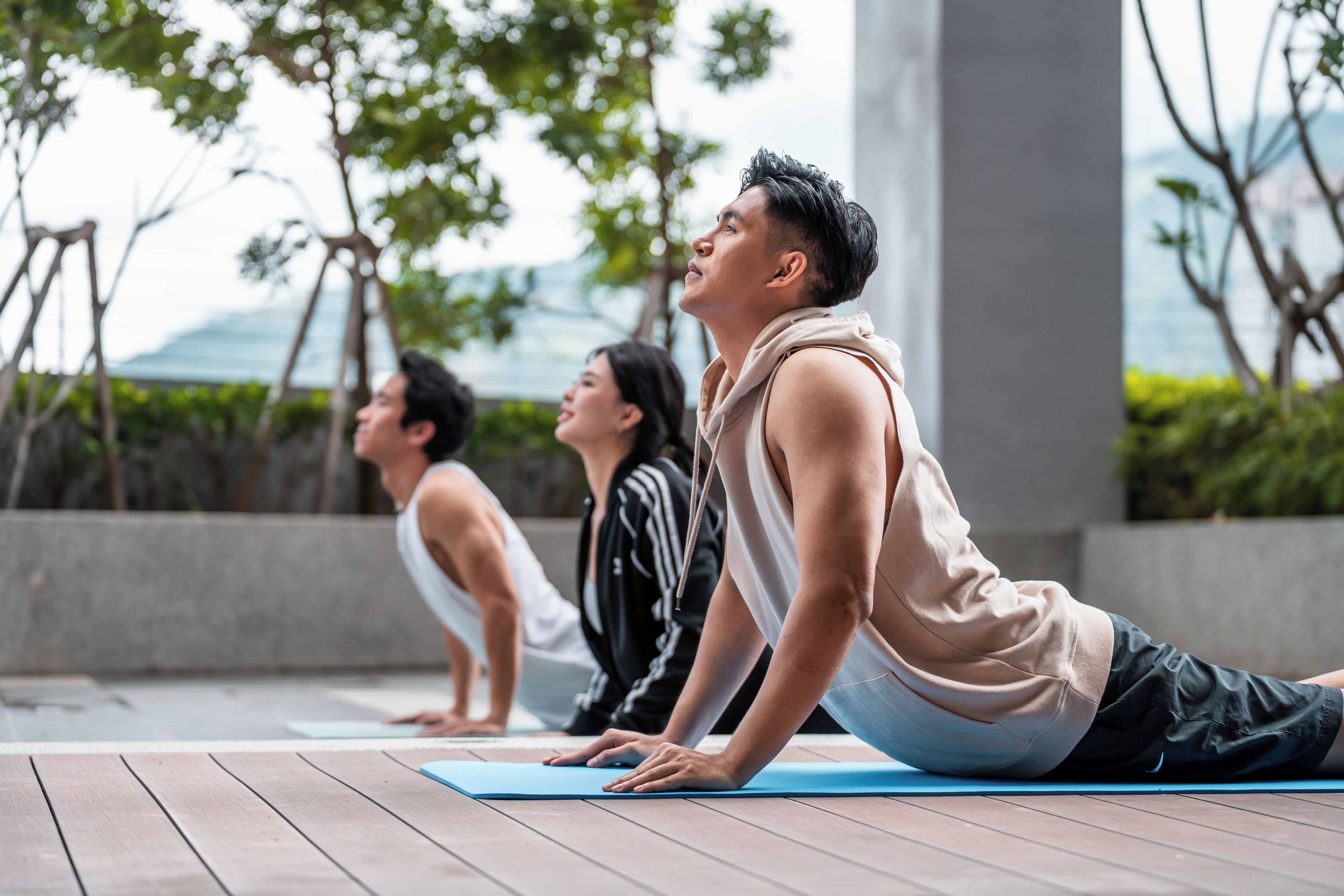 Three individuals performing yoga on a wooden deck with a view of trees and a building in the background.