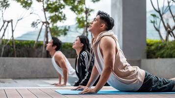 Three individuals performing yoga on a wooden deck with a view of trees and a building in the background.