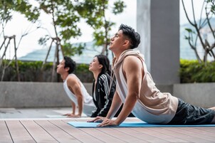 Tres personas practicando yoga en una terraza de madera, con vistas a árboles y un edificio al fondo.