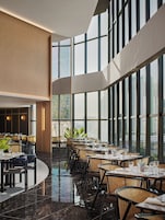 Un restaurante moderno con techos altos y amplias ventanas, que presenta un suelo de mármol pulido y diversas opciones de asientos.