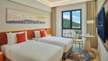 Habitación de hotel con dos camas, un cuadro de un paisaje urbano, un ventanal con vistas, una mesita y un televisor.