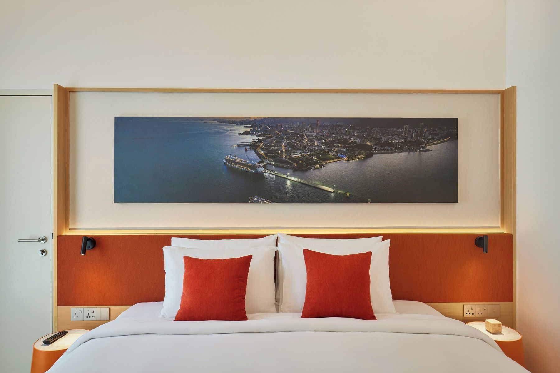 A hotel room with a large framed cityscape artwork above the bed, a wooden headboard, and two red pillows.