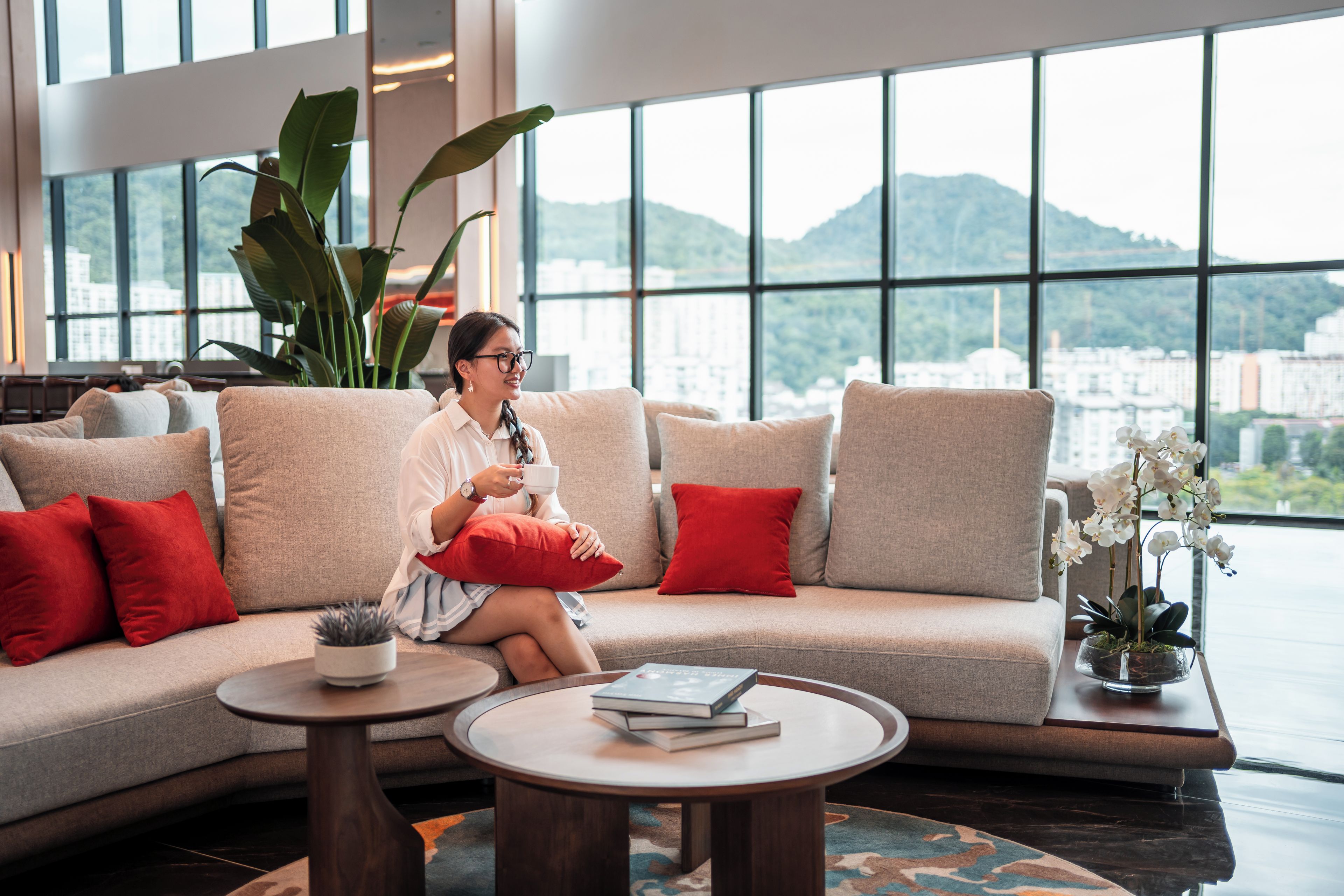 Eine Person sitzt auf einem Sofa und blickt durch große Fenster auf eine Stadtlandschaft.