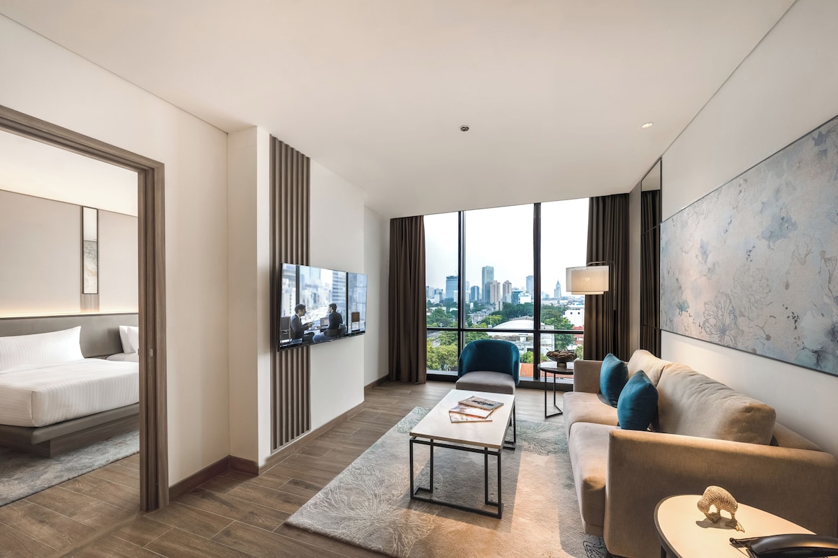 Une chambre d’hôtel moderne équipée d’un lit, d’un canapé, d’une table basse, d’une télévision et d’une grande fenêtre donnant sur la ville.